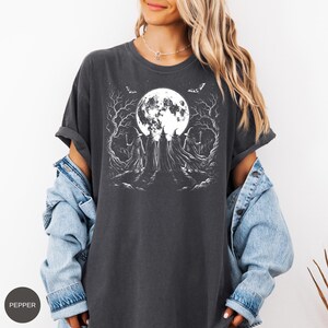 May include: A charcoal gray short-sleeve t-shirt featuring a white graphic print of four cloaked figures in pointed hats standing before a large full moon, surrounded by bare trees and flying bats. The shirt has a crew neckline and rolled sleeves.