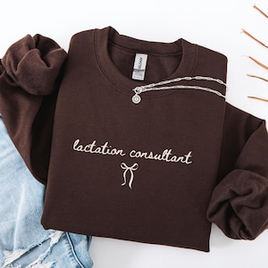 May include: A dark brown sweatshirt with the words "lactation consultant" embroidered in a cursive font, along with a bow graphic. A silver necklace with a pendant is draped over the sweatshirt. The sweatshirt is folded on a white surface.