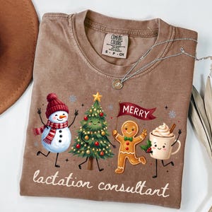 May include: A brown t-shirt featuring a graphic of a snowman, a decorated Christmas tree, a gingerbread person holding a red flag with the text MERRY, and a mug of hot cocoa. The text lactation consultant is printed at the bottom.