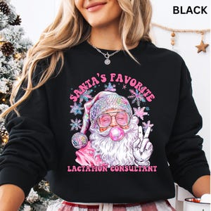 May include: Black sweatshirt with a Santa Claus graphic. Santa is wearing a pink hat, glasses, and blowing a bubblegum bubble. The text reads "SANTA'S FAVORITE LACTATION CONSULTANT" in pink and white.