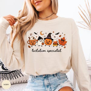 May include: An ivory long-sleeve sweatshirt featuring a graphic print of a coffee mug, a ghost, a jack-o'-lantern, a candy corn, and an autumn leaf with faces. The text "lactation specialist" is printed in black cursive below the characters.