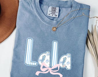 May include: A blue washed denim t-shirt with the name "Lala" in a playful font. The "L" and "a" have blue and white stripes, while the other "a" has a floral pattern. A pink bow is below the name. A gold necklace is draped across the shirt.