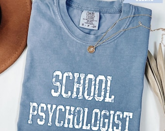 May include: A light blue t-shirt with a white floral design and the text "SCHOOL PSYCHOLOGIST" printed on the front.