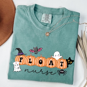 May include: A teal Comfort Colors t-shirt with a Halloween-themed graphic. The design includes pumpkins spelling "FLOAT", a witch hat, bat, ghost, black cat, spider, and the word "nurse". A silver necklace is draped across the shirt.