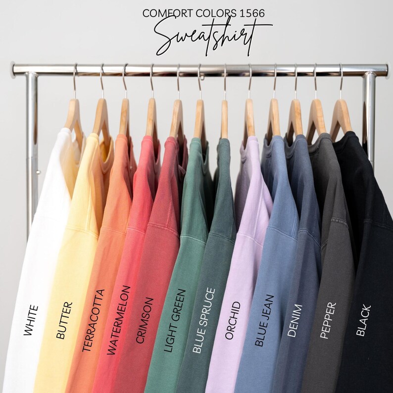 May include: A rack of 12 sweatshirts in various colors, including white, butter, terracotta, watermelon, crimson, light green, blue spruce, orchid, blue jean, denim, pepper, and black. The rack is labeled "COMFORT COLORS 1566 Sweatshirt".