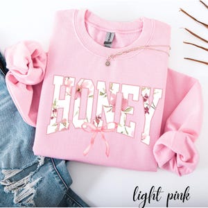 May include: Light pink sweatshirt with the word "HONEY" in white block letters filled with a floral pattern. A pink bow is below the word. A gold necklace with a small pendant is on the sweatshirt. The words "light pink" are at the bottom.