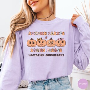 May include: A light purple long-sleeve shirt with the text "AUTUMN LEAVES & BABIES PLEASE LACTATION CONSULTANT". The shirt features four smiling cartoon pumpkins in orange with different accessories.