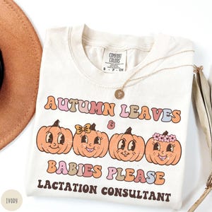 May include: Ivory-colored t-shirt with the text "AUTUMN LEAVES & BABIES PLEASE LACTATION CONSULTANT" in retro font. The shirt features four smiling pumpkin graphics. A gold necklace and a brown hat are also visible.