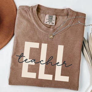 May include: A brown short-sleeve t-shirt with a crew neckline. The shirt features large, cream-colored block letters spelling 'ELL' with the word 'teacher' written in a dark blue cursive script layered over the letters. A silver necklace rests on the shirt.