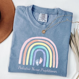 May include: Blue t-shirt with a rainbow graphic and the text "Pediatric Nurse Practitioner". The rainbow features pastel colors, and the design includes baby footprints. A gold necklace rests on the shirt.