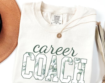 May include: Cream-coloured t-shirt with the words "career COACH" in green block letters filled with a leafy pattern. A gold necklace with a pendant rests on the shirt. A brown hat and dried leaves are in the background.