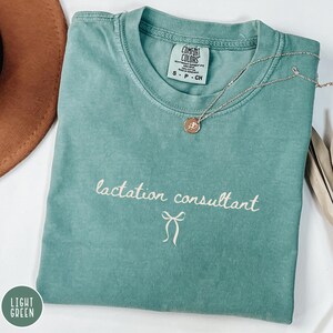 May include: A light green t-shirt with the words "lactation consultant" in cursive script and a bow graphic. A silver necklace with a pendant rests on the shirt. The shirt has a Comfort Colors tag.