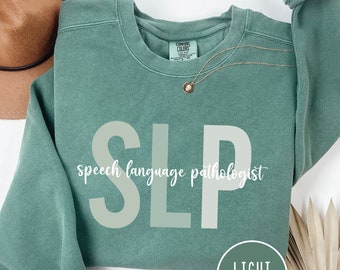May include: A light green sweatshirt with the text "SLP speech language pathologist" in white and light green letters. The sweatshirt is on a white background with a brown hat and a green plant.
