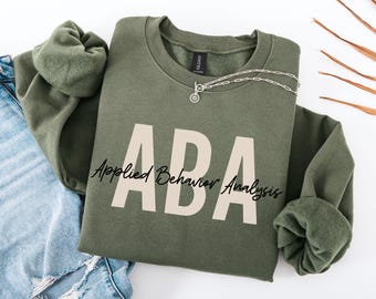 May include: An olive green sweatshirt with the letters "ABA" in large, light beige block letters. Below the letters is the text "Applied Behavior Analysis" in a cursive font. A silver necklace with a circular pendant rests on the sweatshirt.