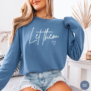 May include: A long-sleeved, blue jean-colored sweatshirt with the words "Let them" in white script and a small heart. The sweatshirt has a crew neck and is made of a soft material. The image also shows a pair of denim shorts.