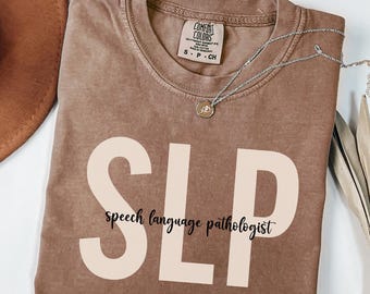 May include: A brown t-shirt with the text "SLP speech language pathologist" printed in white on the front. The shirt is folded and laid flat on a white surface.