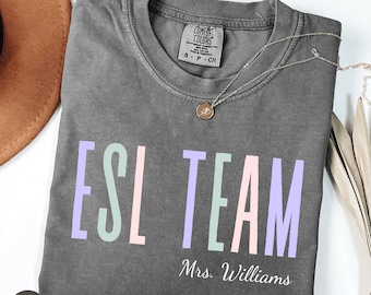 May include: A charcoal gray short-sleeve t-shirt with the text ESL TEAM printed in large, vertical, pastel-colored letters. Below the text, the name Mrs. Williams is printed in white script. The shirt features a Comfort Colors brand neck label.