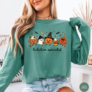 May include: A teal long-sleeve sweatshirt featuring a graphic print of a smiling coffee mug, a ghost, a jack-o'-lantern wearing a witch hat, a candy corn, and an autumn leaf. The text "lactation specialist" is printed in black cursive below.