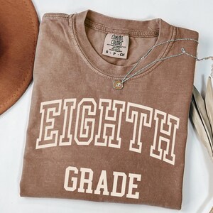 May include: A brown Comfort Colors t-shirt with the words "EIGHTH GRADE" in a white, collegiate-style font. A silver necklace with a small pendant is draped across the shirt. The shirt is folded and lies on a white surface.