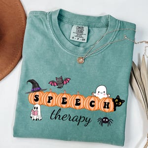May include: A teal Comfort Colors t-shirt with a Halloween-themed design. The design includes pumpkins spelling "SPEECH", a witch hat, a bat, a ghost, a black cat, a spider, and the word "therapy". A silver necklace is draped across the shirt.