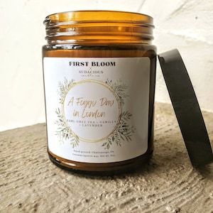 May include: A brown glass candle jar with a gold lid. The label reads "First Bloom x Audacious Candle Co. A Foggy Day in London Earl Grey Tea - Vanilla - Lavender Hand-poured. Chattanooga, TN. Coconut apricot wax. Net wt. 7oz."