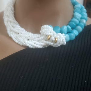 Könnte beinhalten: Eine weiße und türkisfarbene Perlenkette mit einem goldenen Verschluss. Die Kette besteht aus kleinen, runden Perlen und wird um den Hals getragen.