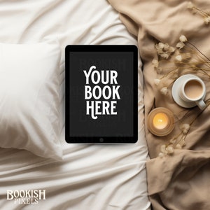 Puede incluir: Una tableta negra con un fondo blanco que dice "Your Book Here" en la pantalla. La tableta está sobre una cama blanca con una manta marrón y una vela encendida y una taza de café sobre la manta.