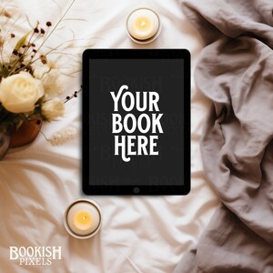 Puede incluir: Una tableta negra con una pantalla blanca que muestra el texto "YOUR BOOK HERE" en una cama blanca con dos velas encendidas y un ramo de flores blancas.
