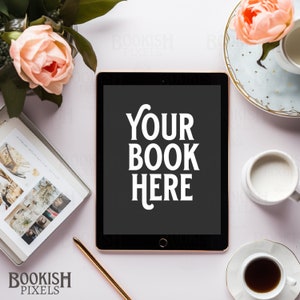 Puede incluir: Una tableta negra con una pantalla blanca que muestra el texto "YOUR BOOK HERE". La tableta está sobre una superficie blanca con una taza de café, un plato de galletas y un ramo de flores rosas.