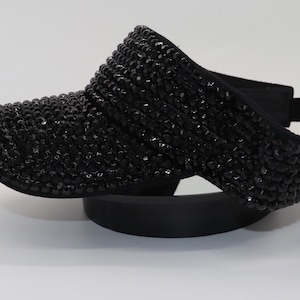 May include: A black visor with a black rhinestone design. The visor is made of a soft, flexible material and has a curved brim.