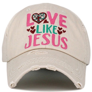 May include: A beige distressed baseball cap with the text "Love Like Jesus" embroidered in pink, green, and red. The text includes a heart with a cross inside.