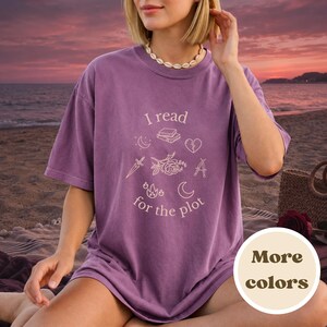 May include: A purple short-sleeve t-shirt featuring the text I read for the plot in white, accompanied by line art illustrations of books, a heart, a dagger, a rose, flames, and moons. The shirt has a crew neckline and a relaxed fit.