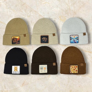 Könnte beinhalten: Sechs gerippte Strickmützen in verschiedenen Farben: Beige, Creme, Hellgrau, Schwarz, Braun und Dunkelbraun. Jede Mütze hat einen quadratischen Aufnäher mit verschiedenen Designs, darunter ein Bär, ein Tiger, eine Landschaft, eine Katze, Kaffeetassen und Herbstblätter.