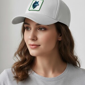 Puede incluir: Gorra de b&eacute;isbol gris claro con un parche que muestra una ilustraci&oacute;n de un lobo en tonos azules y verdes, enmarcada por un borde verde. La gorra tiene una visera curva y una corona estructurada.