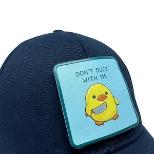 Pode incluir: Um boné de beisebol azul-marinho com um emblema quadrado azul-claro na frente. O emblema mostra um pato amarelo de desenho animado segurando uma faca prateada com o texto "DON'T DUCK WITH ME" impresso em letras azul-escuras.