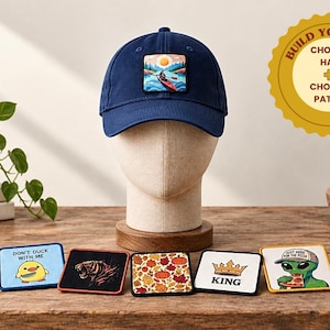 May include: A navy blue baseball cap featuring a rectangular patch of a person kayaking on a lake at sunset. Five additional rectangular patches are displayed in front, featuring a duck, a tiger, autumn leaves, a crown, and an alien eating pizza.