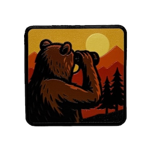 May include: A square patch featuring a brown bear looking through binoculars at a sunset over mountains and pine trees. The design uses a warm color palette of yellow, orange, and deep red, with a black border framing the entire graphic illustration.