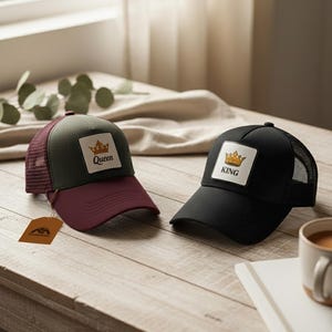 Könnte beinhalten: Zwei Trucker-Caps mit Kronenaufnähern. Eine Kappe ist schwarz mit dem Text "KING", die andere ist grün und weinrot mit dem Text "QUEEN". Beide haben Mesh-Rücken und liegen auf einem Holztisch.