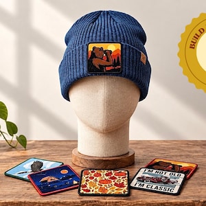 May include: A navy blue ribbed knit beanie featuring a square patch of a bear drinking from a mug against a sunset landscape. The beanie is displayed on a mannequin head, accompanied by several square patches with various graphic designs.