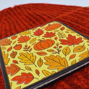 May include: A square coaster featuring a yellow background with a repeating pattern of orange pumpkins, red maple leaves, yellow oak leaves, and small brown acorns. The coaster has a thick black border and a layered edge, resting on a textured red surface.