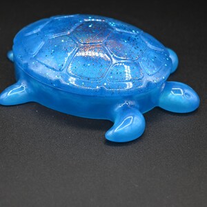 May include: A translucent blue turtle-shaped trinket box with a smooth, glossy finish. The shell features a moulded hexagonal pattern embedded with fine, shimmering glitter particles that catch the light, creating a subtle sparkle across the surface.