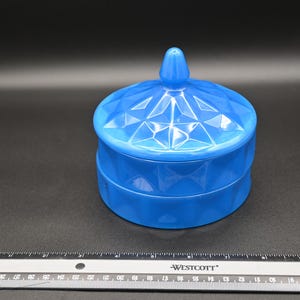 May include: A vibrant blue, lidded trinket box with a geometric pattern. The round box has a matching lid with a small, pointed finial. The box is made of a glossy material and sits on a dark surface.