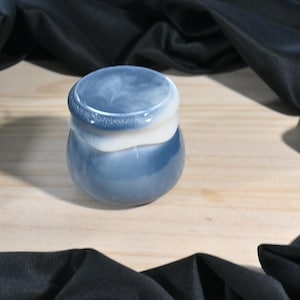 Op de afbeelding: Een kleine, ronde, blauw en witte keramische pot met een deksel. De pot heeft een glanzende afwerking en een gemarmerd uiterlijk. De pot staat op een lichtgekleurd houten oppervlak, met zwarte stof eromheen.