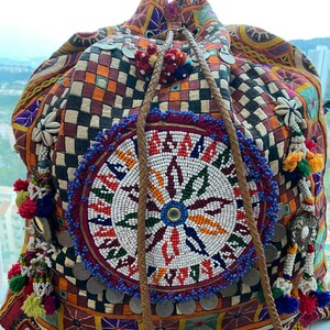 Può includere: Una colorata borsa a coulisse fantasia con un design rotondo di perline. La borsa presenta un mix di quadrati arancioni, rossi e blu, con nappe decorative, monete e un cinturino in pelle. Il design centrale è un motivo circolare di perline bianche, rosse e blu.