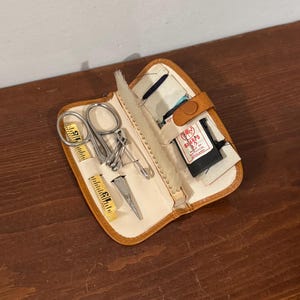 May include: A small, open sewing kit with a tan leather exterior and cream-coloured interior. The kit contains small scissors, a tape measure, needles, and other sewing tools. The kit is open, displaying its contents.