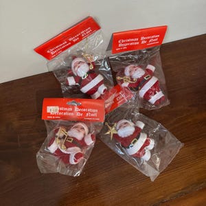 Può includere: Diverse decorazioni natalizie, ognuna raffigurante una piccola figura di Babbo Natale in abito rosso con finiture bianche, confezionate in plastica trasparente. Le confezioni includono etichette rosse con la scritta "Christmas Decoration Decoration De Noel".