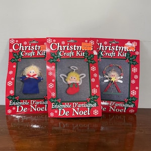 May include: Three Christmas craft kits with red packaging, featuring holly and snowflake designs. Each kit contains a felt ornament: an angel in a blue robe, an angel in a red stocking, and a star-shaped angel. The text "Christmas Craft Kit" is visible.