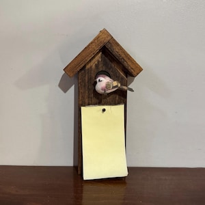 Può includere: Una casetta per uccelli in legno con un piccolo uccello decorativo all'ingresso. La casetta ha un tetto a capanna e un blocco note rettangolare attaccato sul davanti. L'uccello è rosa, marrone e bianco. Il blocco note è giallo.