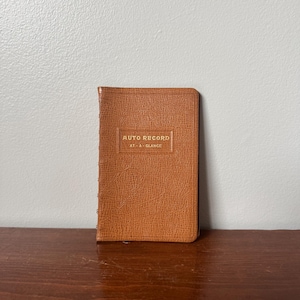 Peut inclure: Un carnet rectangulaire avec une couverture beige texturée. La couverture avant présente le texte doré en relief "AUTO RECORD" au-dessus des mots "AT-A-GLANCE" encadrés par un rectangle. Le carnet possède une reliure verticale sur le côté gauche.