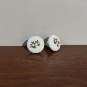 Peut inclure: Deux boutons de meuble en céramique blanche avec un motif floral. Chaque bouton est de forme ronde avec un groupe de fleurs peintes en rose, bleu et rouge, avec des feuilles vertes. Les boutons sont posés sur une surface marron foncé.